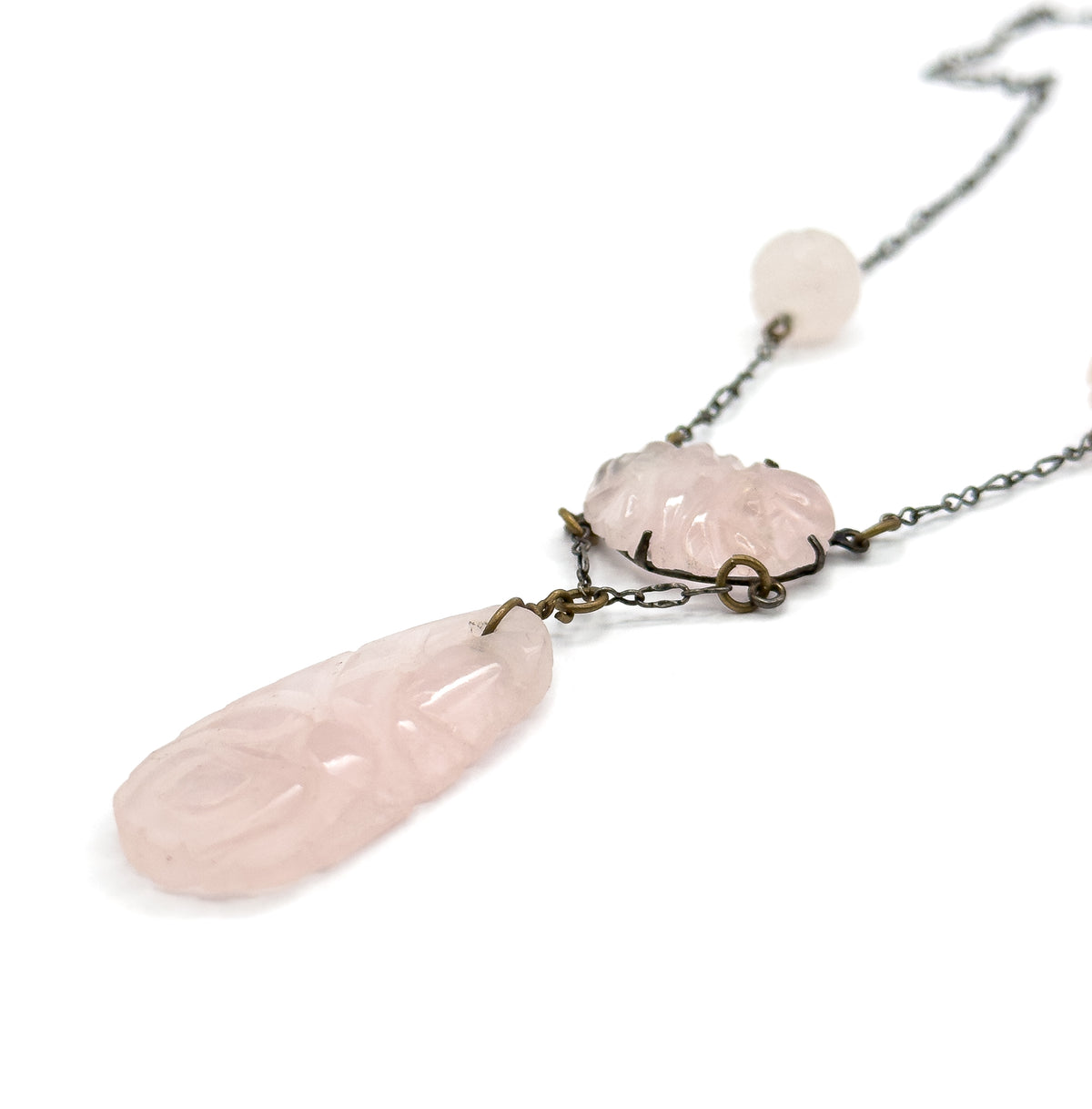 Close angled view of the lower half on white, the long carved rose quartz drop swinging from its jump ring beneath the pronged oval centre station, with a round carved bead set into the chain above.