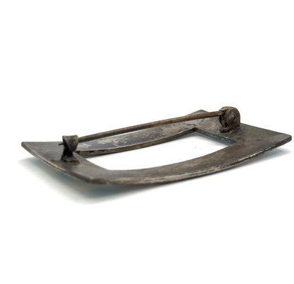 A low angled view across the back of the brooch, showing the darkened rectangular frame edge-on with the pin stem raised above it between hinge and catch.
