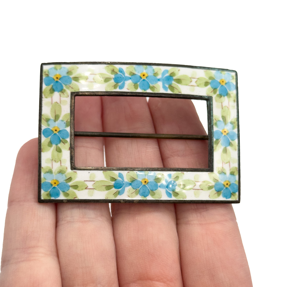 Shown face-up resting on open fingers for scale, the open rectangular frame painted with blue and yellow flowers and green leaves on white enamel.