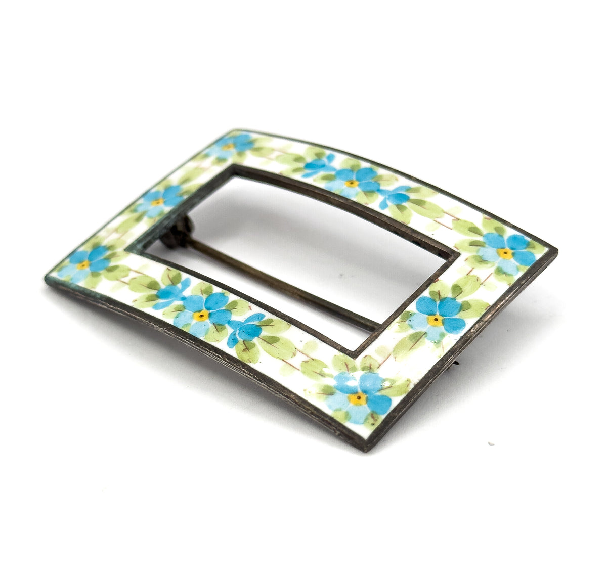Angled front view from one corner, the white enamel band and its blue and yellow flowers curving away in perspective with the pin stem glimpsed through the open centre.