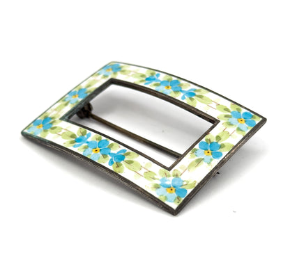 Angled front view from one corner, the white enamel band and its blue and yellow flowers curving away in perspective with the pin stem glimpsed through the open centre.