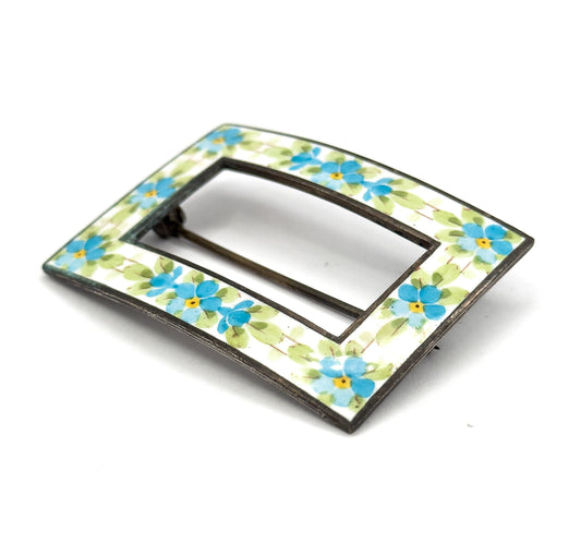 Angled front view from one corner, the white enamel band and its blue and yellow flowers curving away in perspective with the pin stem glimpsed through the open centre.