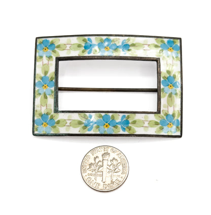 Flat front view with a dime placed below for scale, the white enamel band of blue and yellow flowers framing the open centre with the pin stem crossing it.