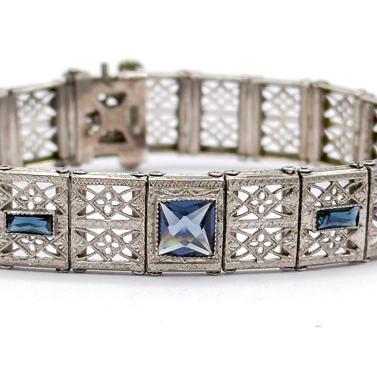 Close in at eye level on three front links, the central square paste catching light to a pale silvery blue between two deep blue rectangles, with the clasp box blurred on the far side behind.