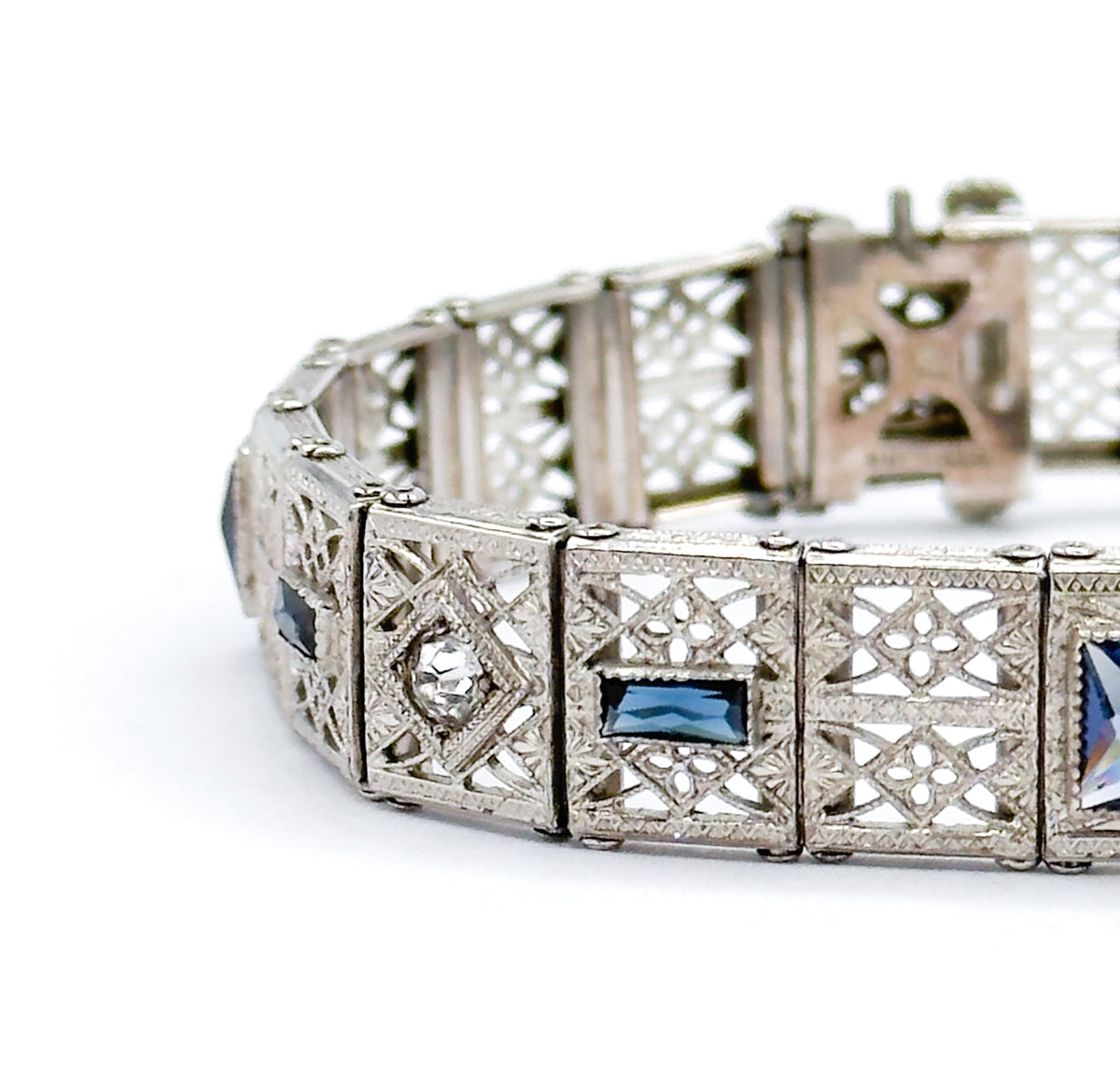 The bracelet curved on a white surface, foreground links in sharp focus showing a clear faceted stone in a square bezel beside a blue paste rectangle, with the clasp end receding behind.