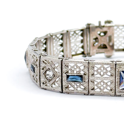 The bracelet curved on a white surface, foreground links in sharp focus showing a clear faceted stone in a square bezel beside a blue paste rectangle, with the clasp end receding behind.