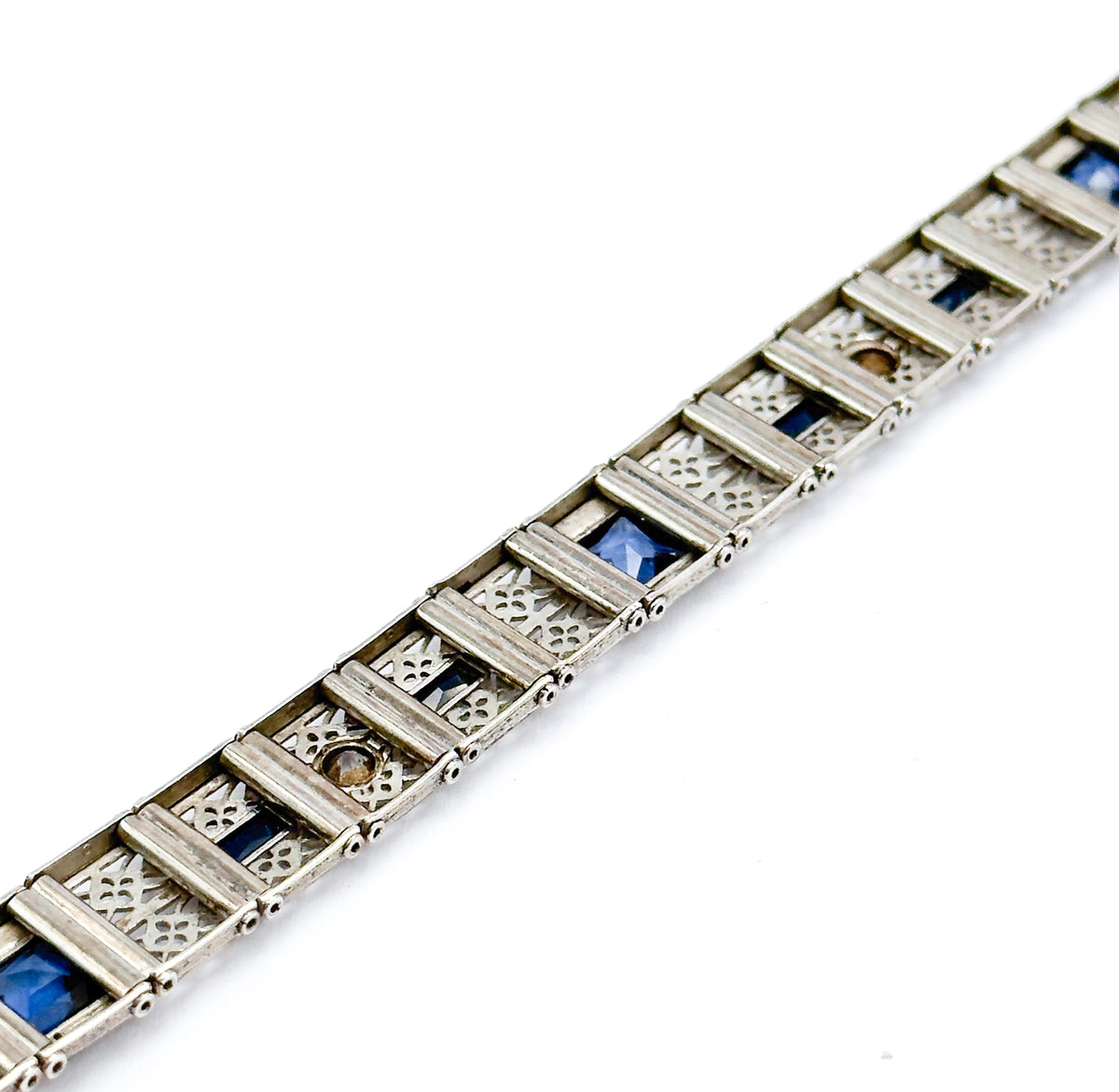 An Art Deco blue paste filigree bracelet in sterling silver, laid flat and angled across a white background, its openwork links alternating with deep blue square stones.
