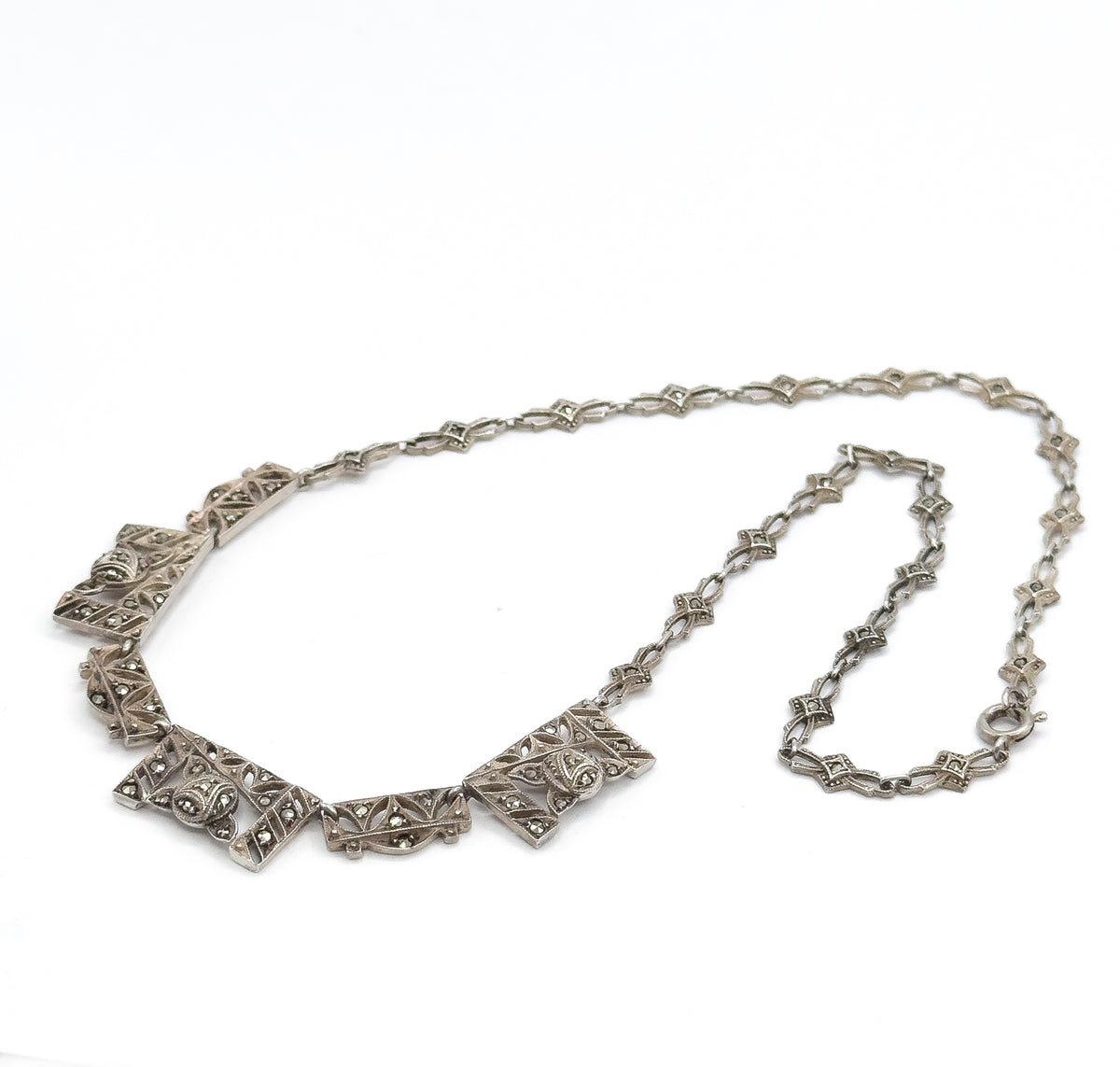 The whole necklace laid open in a loop on white, chevron panels gathered at the lower left and the lozenge-link chain sweeping round to the spring ring clasp at right.