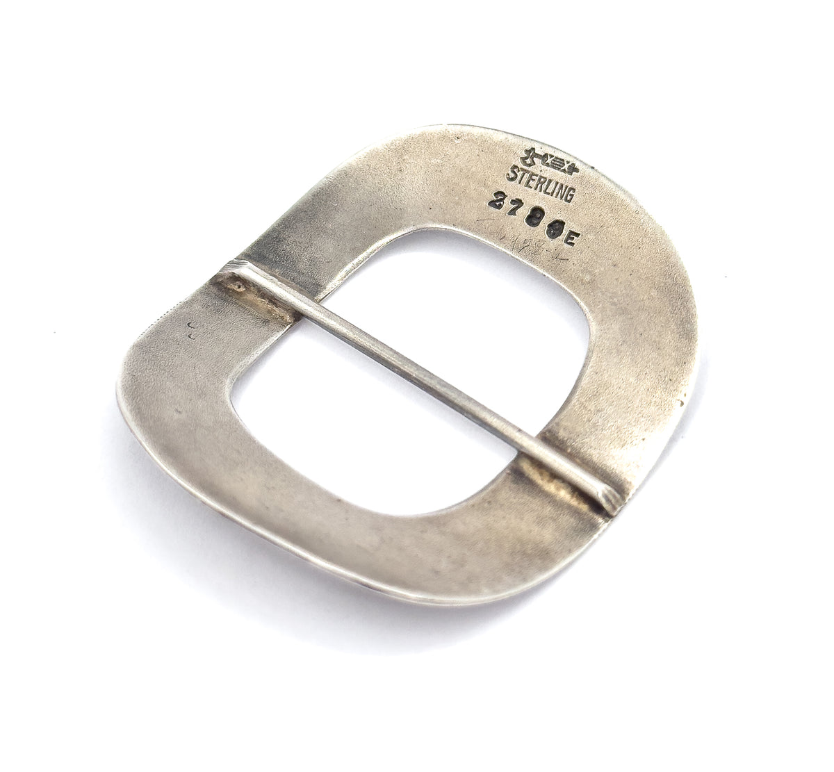 The reverse tilted at an angle on white, catching soft patina across the plain surface and bringing the stamped marks and the soldered ends of the central bar into clearer view.
