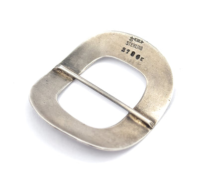 The reverse tilted at an angle on white, catching soft patina across the plain surface and bringing the stamped marks and the soldered ends of the central bar into clearer view.