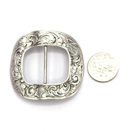 The engraved face flat on white beside a dime for scale, the buckle standing roughly four times the coin's width with its floral scrollwork framing the open centre bar.
