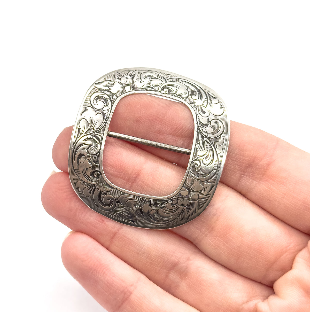 An Edwardian hand engraved floral buckle in sterling silver, shown resting on open fingers that convey its palm-filling size, with a scrolling pattern around the open centre bar.
