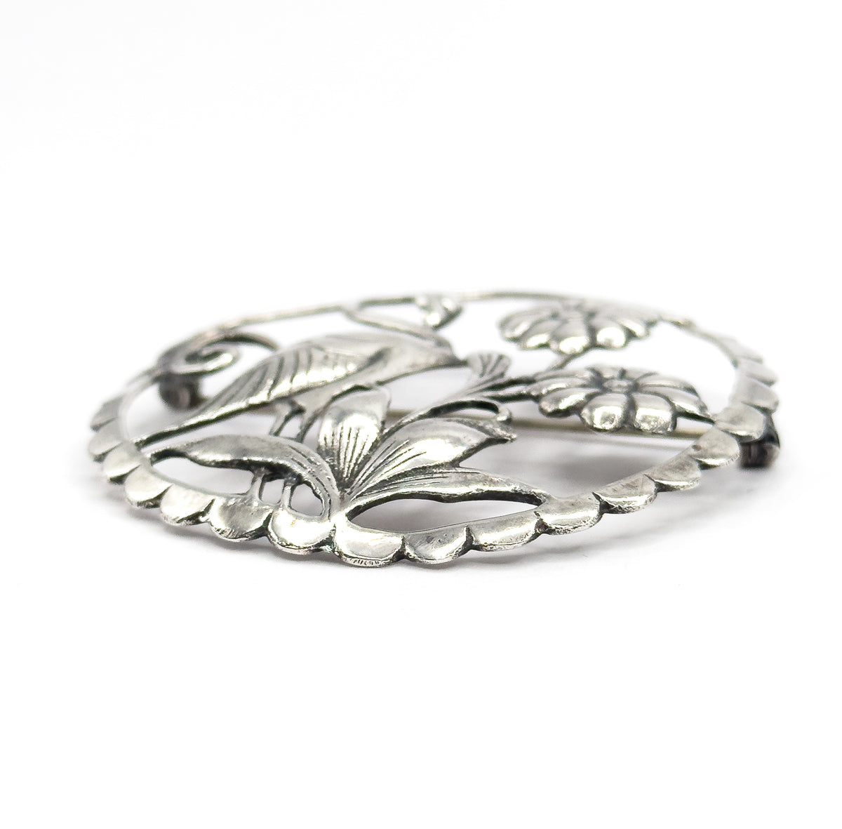 Low three-quarter angle across the brooch, showing how the leaves and flower heads rise in relief above the scalloped rim, with the pin stem glimpsed through the openwork.