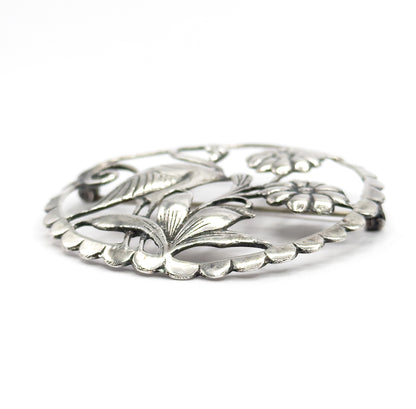 Low three-quarter angle across the brooch, showing how the leaves and flower heads rise in relief above the scalloped rim, with the pin stem glimpsed through the openwork.