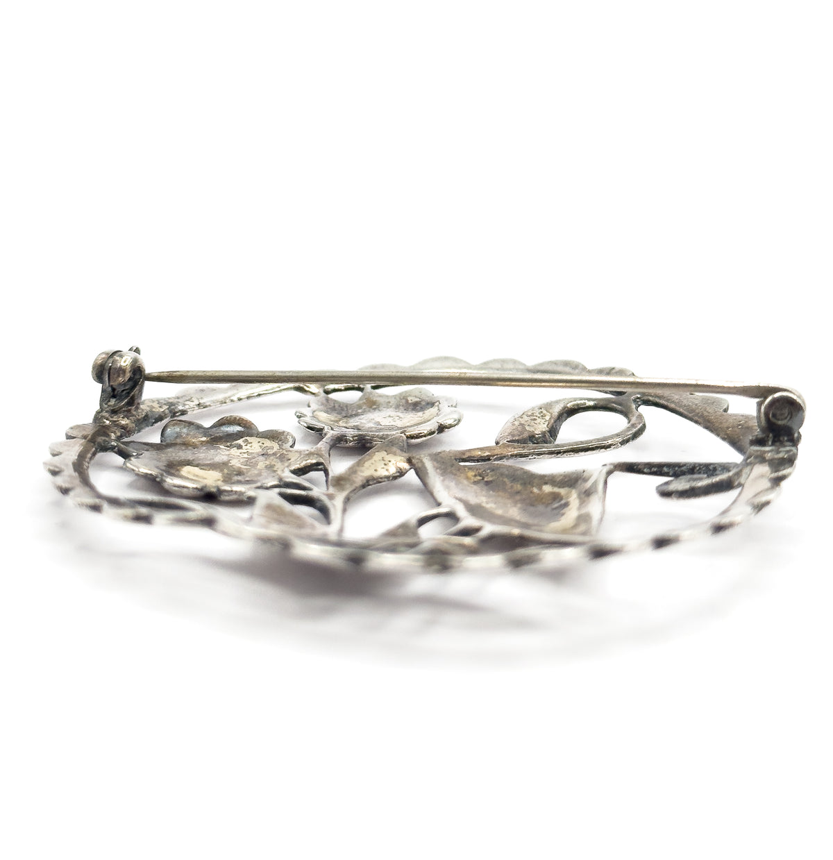 Back of the brooch seen at a low angle, the straight pin stem spanning from its hinge at one side to the C-shaped catch at the other above the hollowed reverse of the flowers.