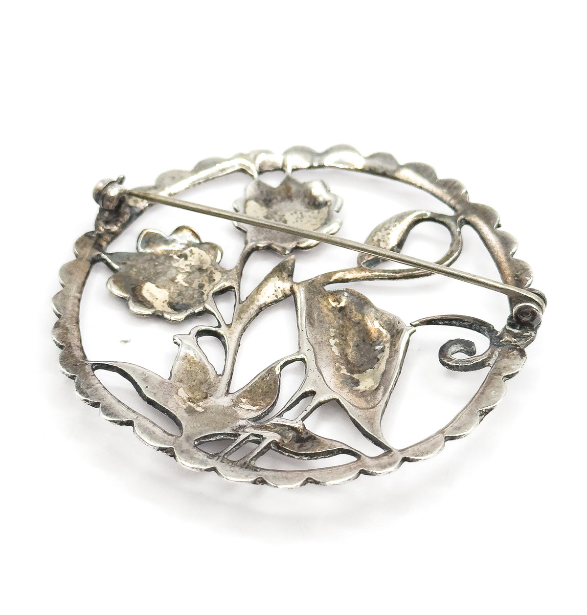 Tilted view of the reverse showing the hollow undersides of the flower heads and leaves, with patina pooled in the recesses and the curled tip of the catch at the right.