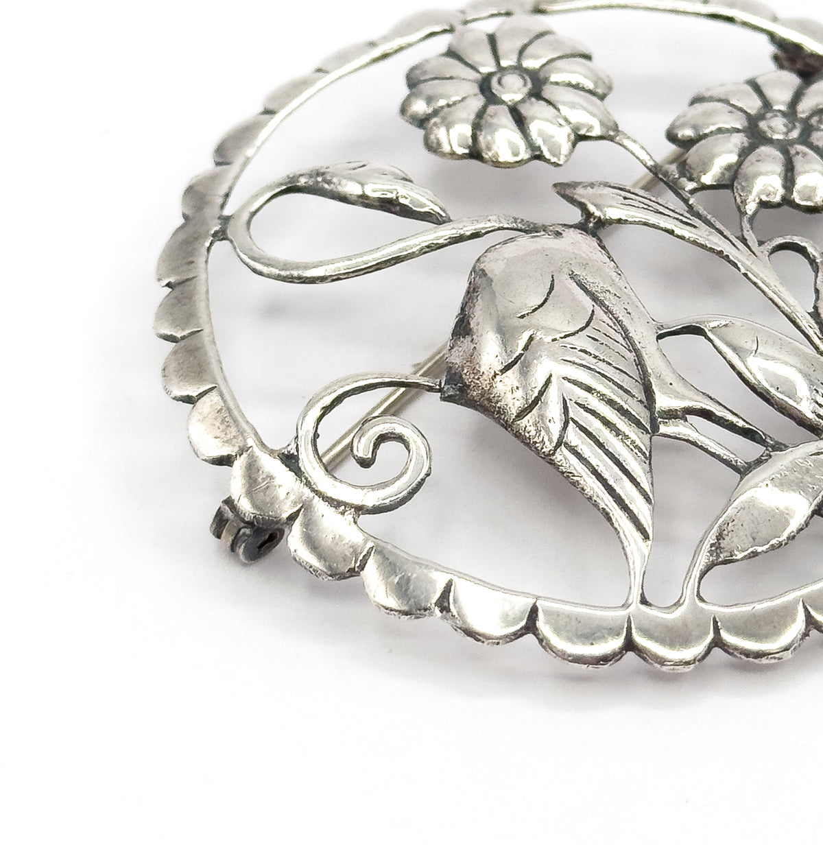 Close crop on one section of the circle, enlarging the scalloped rim, a coiled scroll and a deeply ridged leaf beneath the daisy-like flowers, with the pin stem crossing behind.