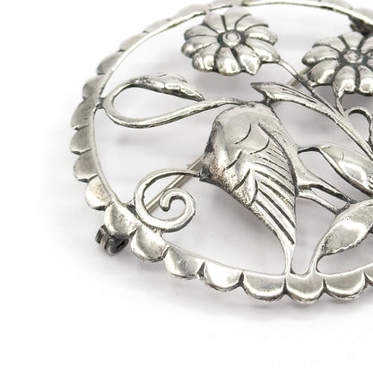 Close crop on one section of the circle, enlarging the scalloped rim, a coiled scroll and a deeply ridged leaf beneath the daisy-like flowers, with the pin stem crossing behind.