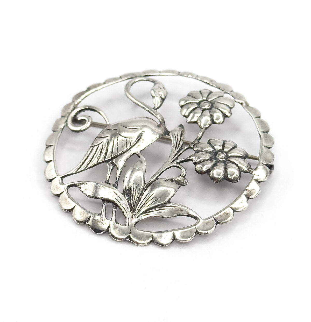 Front of the sterling silver brooch tilted slightly, the long-necked flamingo standing among two round flowers and ridged leaves inside the scalloped circle, pin stem just visible behind.