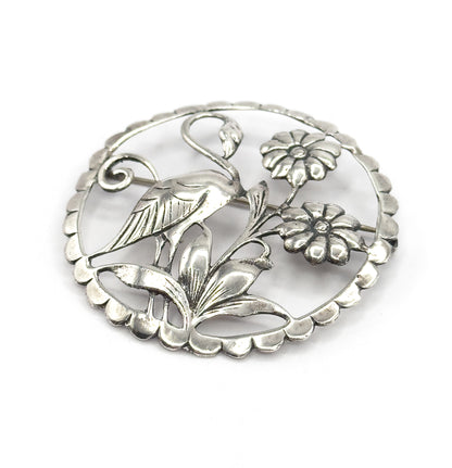 Front of the sterling silver brooch tilted slightly, the long-necked flamingo standing among two round flowers and ridged leaves inside the scalloped circle, pin stem just visible behind.
