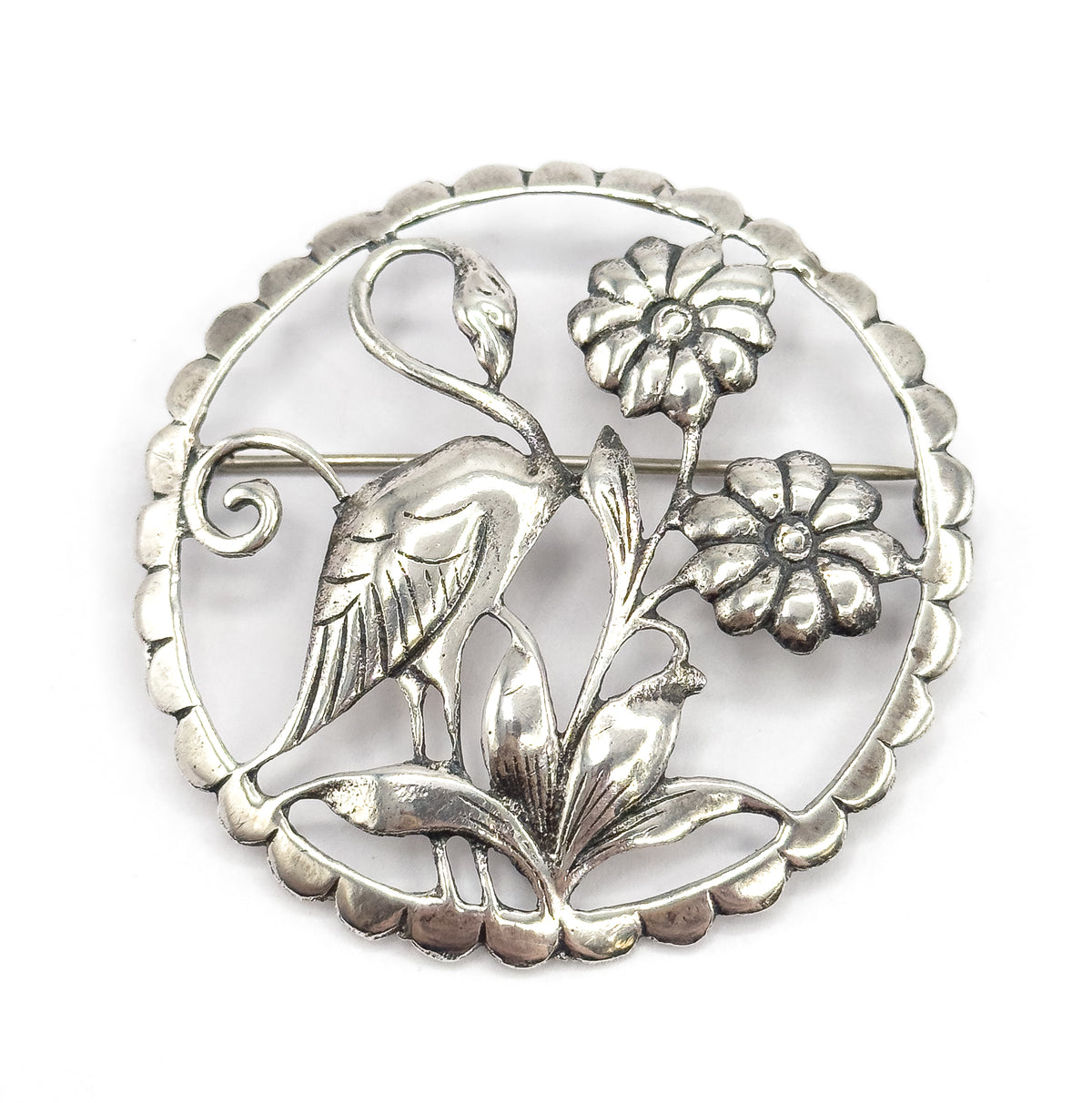 Straight-on front view, the flamingo's curved neck, two round flowers and arching leaves filling the openwork circle, with the pin stem crossing behind the bird's body and patina darkening the recesses.