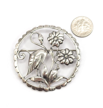 Round sterling silver openwork brooch with a flamingo among flowers inside a scalloped rim, shown flat from the front beside a dime for scale, with the pin stem crossing the back.