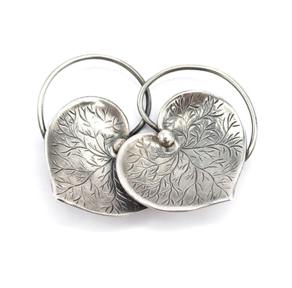 Front view on white with the two wire loops rising high behind like twin arches, the overlapping hearts tipped forward so their leaf-like etched veining radiates out from the single visible centre bead.