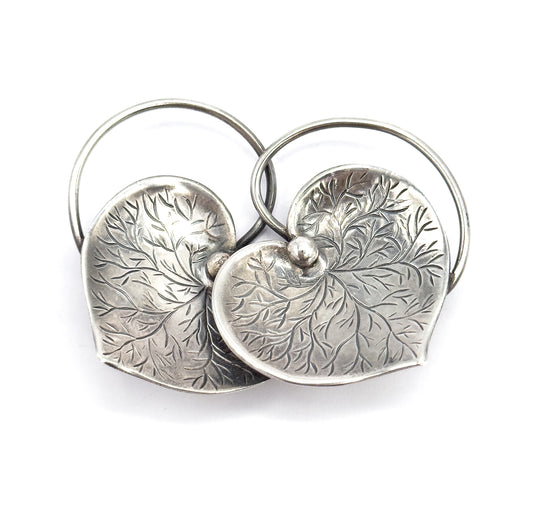 Front view on white with the two wire loops rising high behind like twin arches, the overlapping hearts tipped forward so their leaf-like etched veining radiates out from the single visible centre bead.