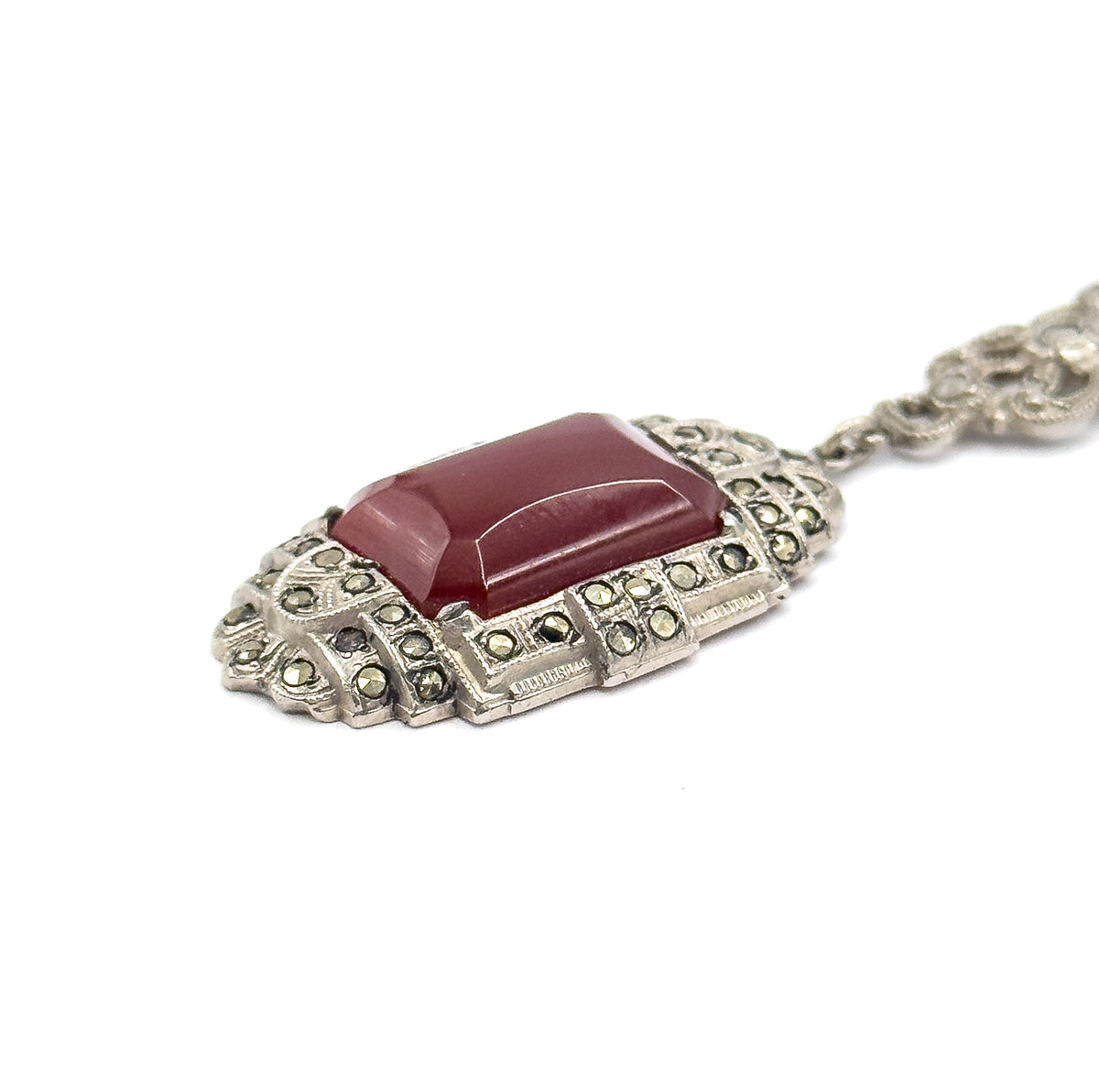 Low, raking close-up along the pendant's front, the carnelian's bevelled edges catching light above tiered rows of marcasites, the bail and one chain station trailing off to the upper right.