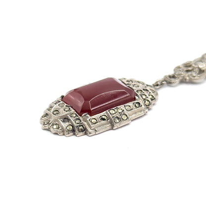 Low, raking close-up along the pendant's front, the carnelian's bevelled edges catching light above tiered rows of marcasites, the bail and one chain station trailing off to the upper right.