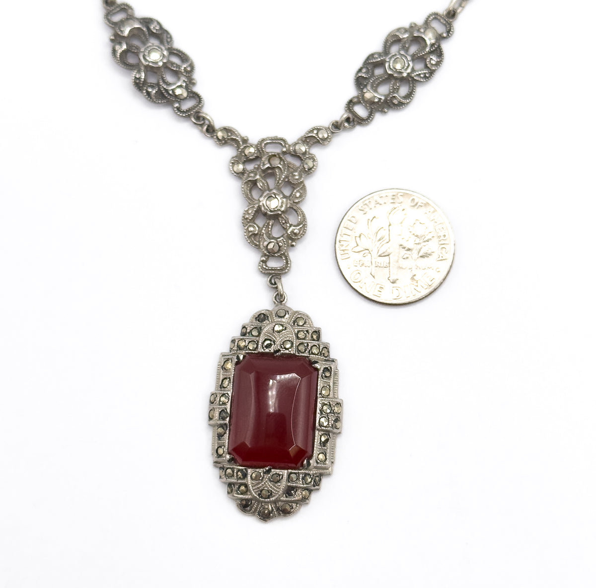 Laid flat beside a dime for scale, the necklace shows its floral openwork marcasite stations, the drop-shaped centre link and the rectangular carnelian pendant hanging below.