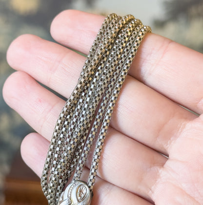 Several strands of the round box chain draped across the fingers of a hand, which gives a sense of its fine scale, with the engraved sliding bead just visible at the lower edge.