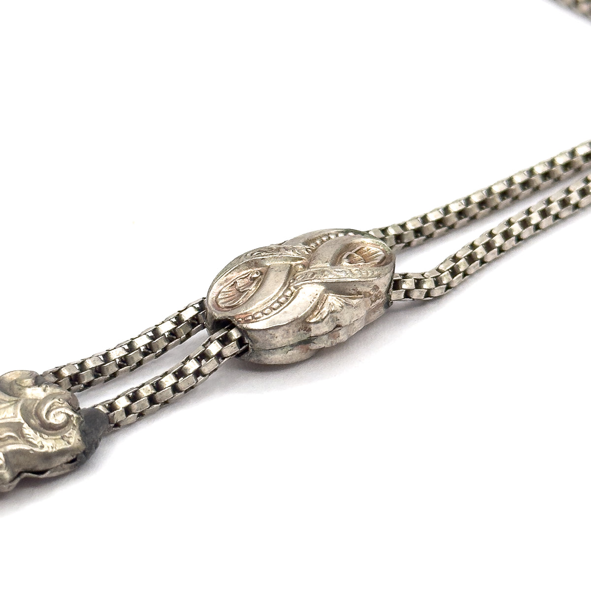 Close view of the engraved sliding bead with the two round box chain strands running through it, and the scrolled fitting at the lower left edge of the frame.