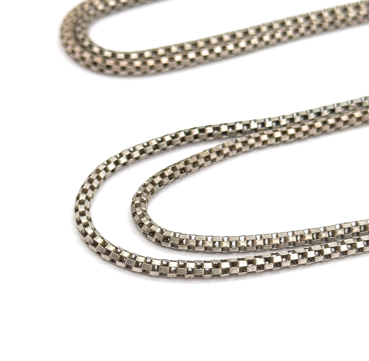 Long looping folds of the round box chain curve back on themselves across plain white, showing the square links of the light-coloured metal with no fittings in frame.