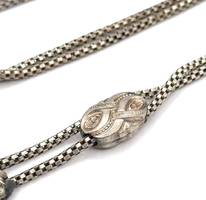 Angled view from above with the engraved sliding bead gathering the two strands into one, further lengths of box chain crossing the upper background and a fitting just entering the lower left corner.