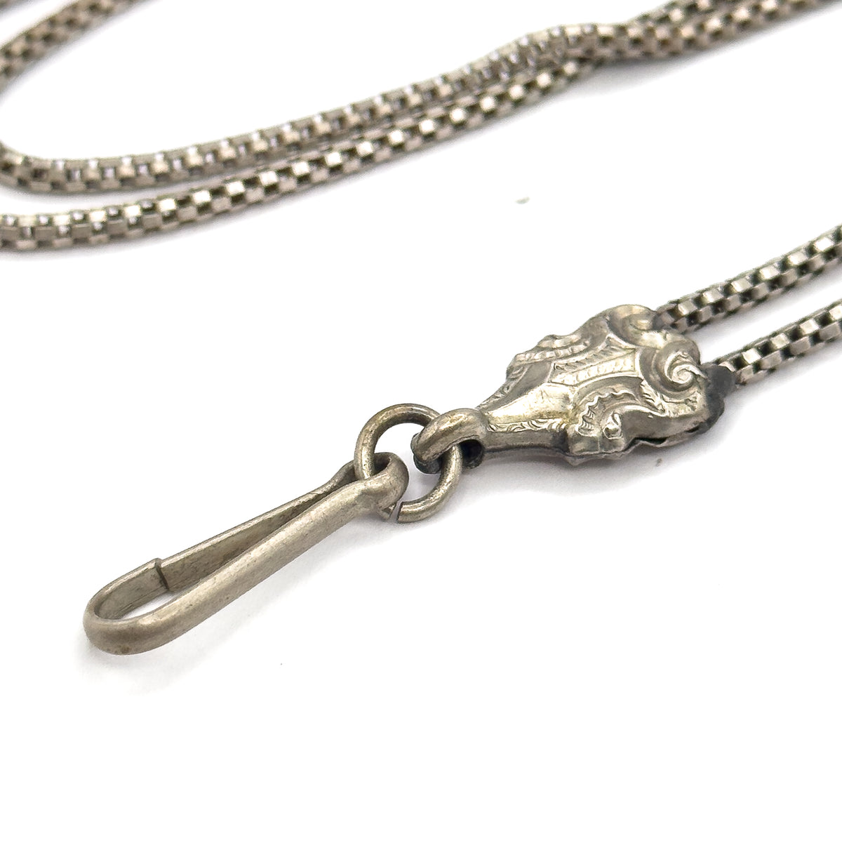 Close view of the long hook clasp hanging from its jump ring beside the scroll-engraved sliding bead, where two strands of box chain pass through and loop away behind.