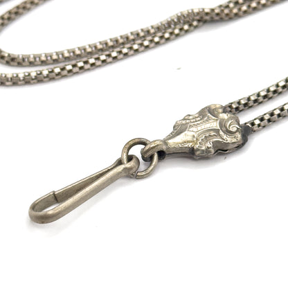 Close view of the long hook clasp hanging from its jump ring beside the scroll-engraved sliding bead, where two strands of box chain pass through and loop away behind.