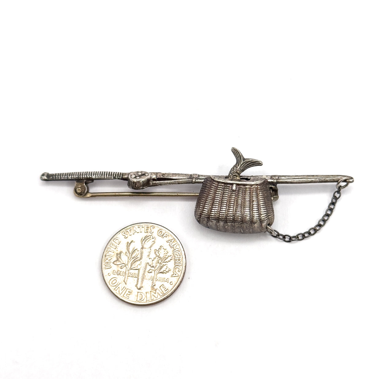 The brooch lies beside a dime for scale, the coin roughly matching the height of the woven basket while the pole, fish tail and safety chain stretch well past it.