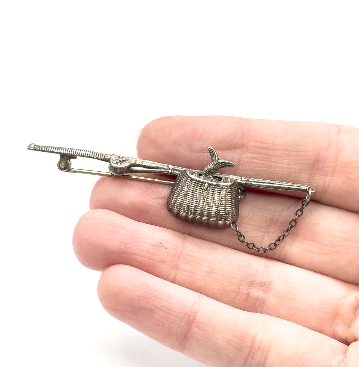Sterling silver fishing pole and basket brooch shown resting across open fingers for scale, with pin stem, safety chain and a small fish tail at the woven basket.