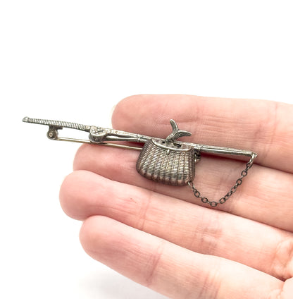 Sterling silver fishing pole and basket brooch shown resting across open fingers for scale, with pin stem, safety chain and a small fish tail at the woven basket.
