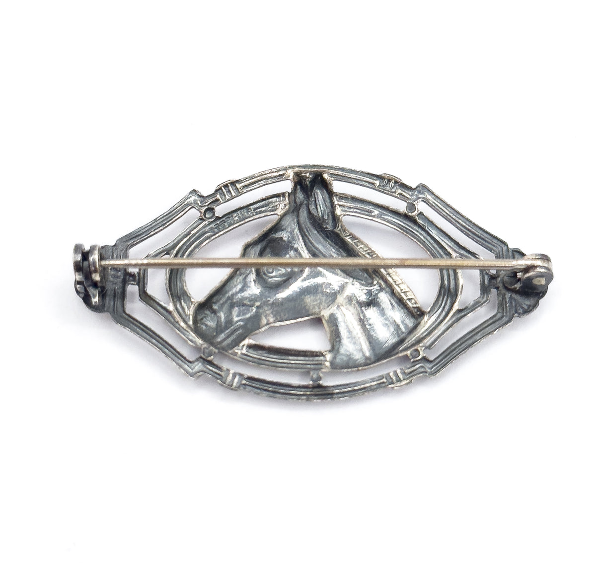 The reverse of the brooch on a white background, showing the straight pin stem crossing the openwork frame between hinge and clasp, with patina pooled behind the horse's head.