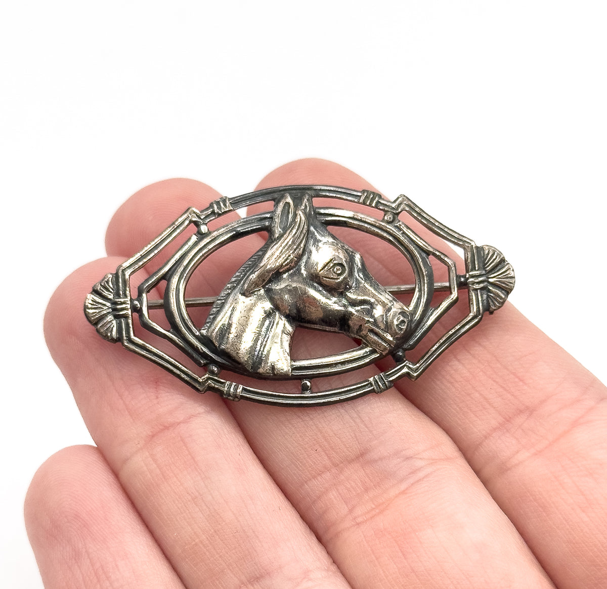 A sterling silver horse head brooch held flat on open fingers, the raised equine profile set within an openwork oval frame, patina darkening the recesses and showing its palm-sized scale.