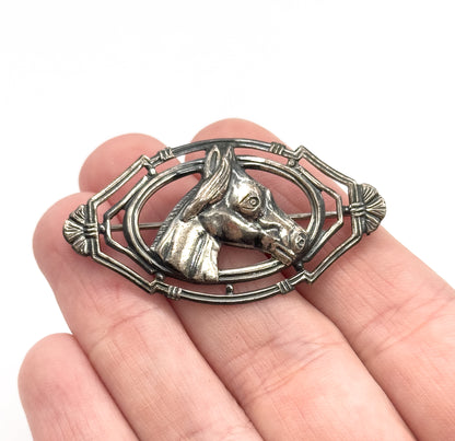 A sterling silver horse head brooch held flat on open fingers, the raised equine profile set within an openwork oval frame, patina darkening the recesses and showing its palm-sized scale.
