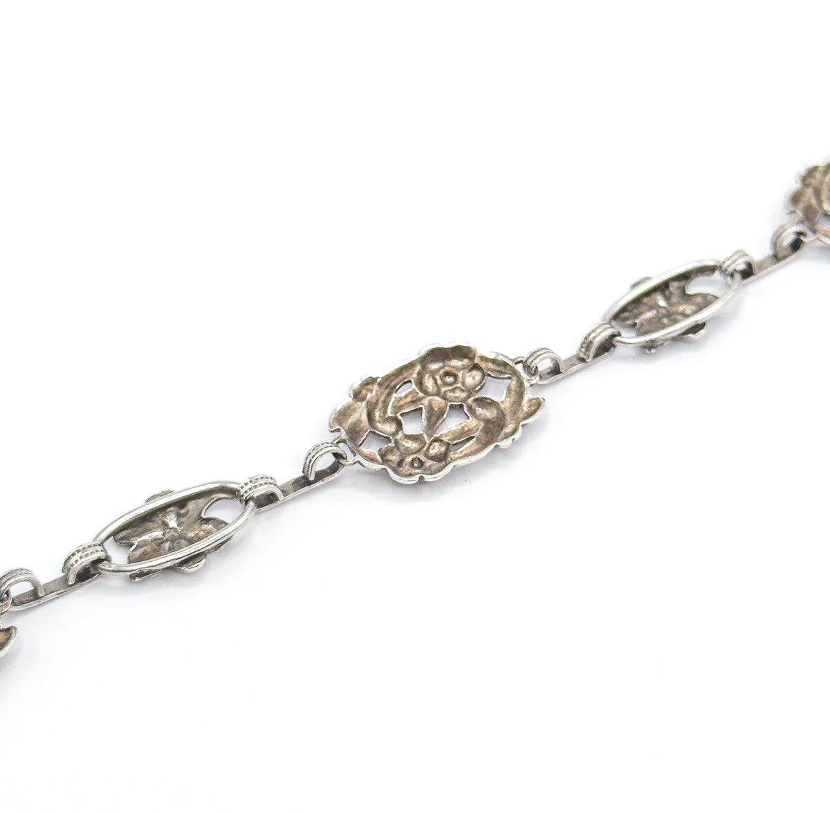 Close diagonal view on a plain white background, showing the chain of alternating pierced oval stations and larger scalloped flower link with patina settled in the openwork.