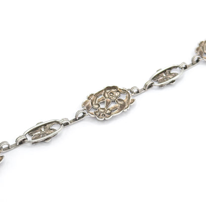 Close diagonal view on a plain white background, showing the chain of alternating pierced oval stations and larger scalloped flower link with patina settled in the openwork.