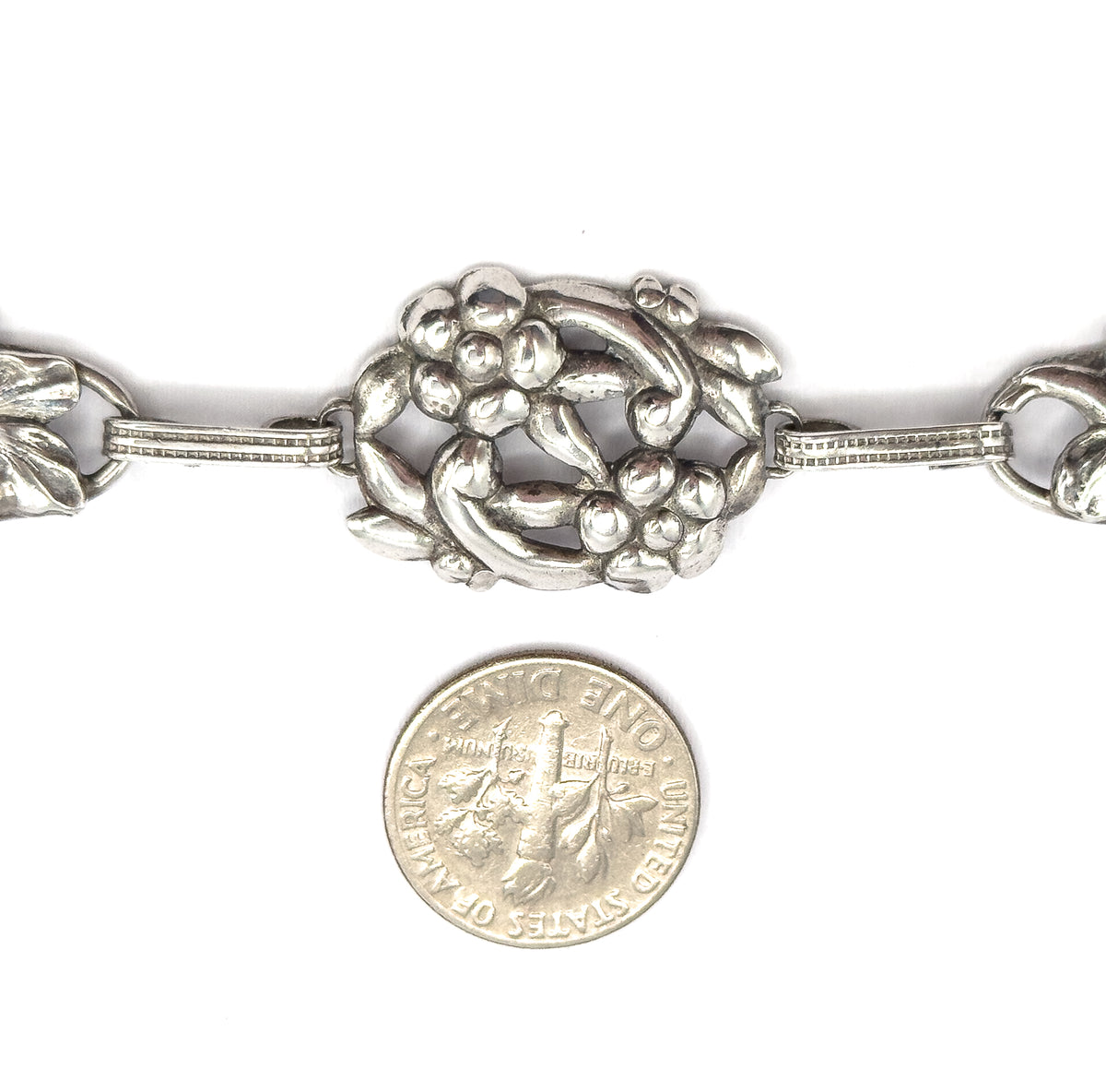 A dime placed below the bracelet gives scale against the central oval floral station and its ribbed bar links, with patina in the recesses.