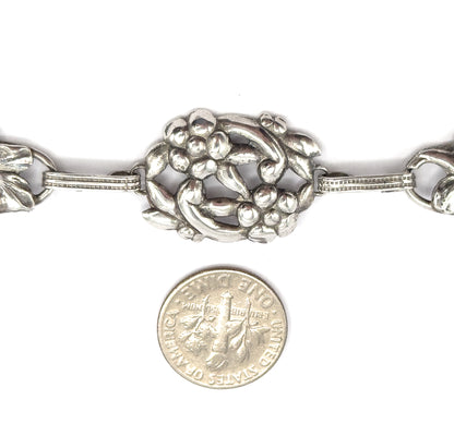 A dime placed below the bracelet gives scale against the central oval floral station and its ribbed bar links, with patina in the recesses.