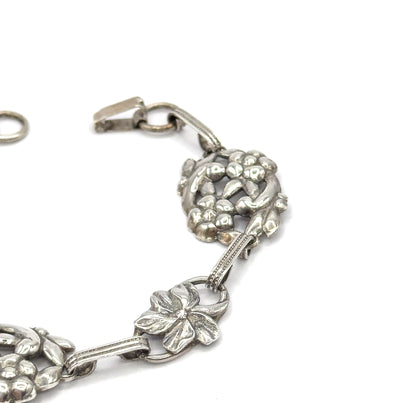 Angled close-up of the unfastened clasp end, hook and round ring apart at the top, beside a domed clustered station and a flatter five-petal flower link below.