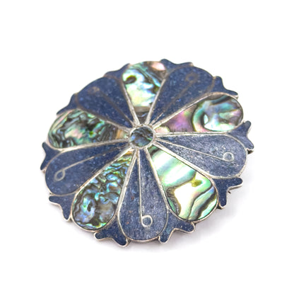 Angled three-quarter view on white showing the domed face and scalloped sterling silver edge, with abalone segments catching green and pink light between the crushed stone wedges.