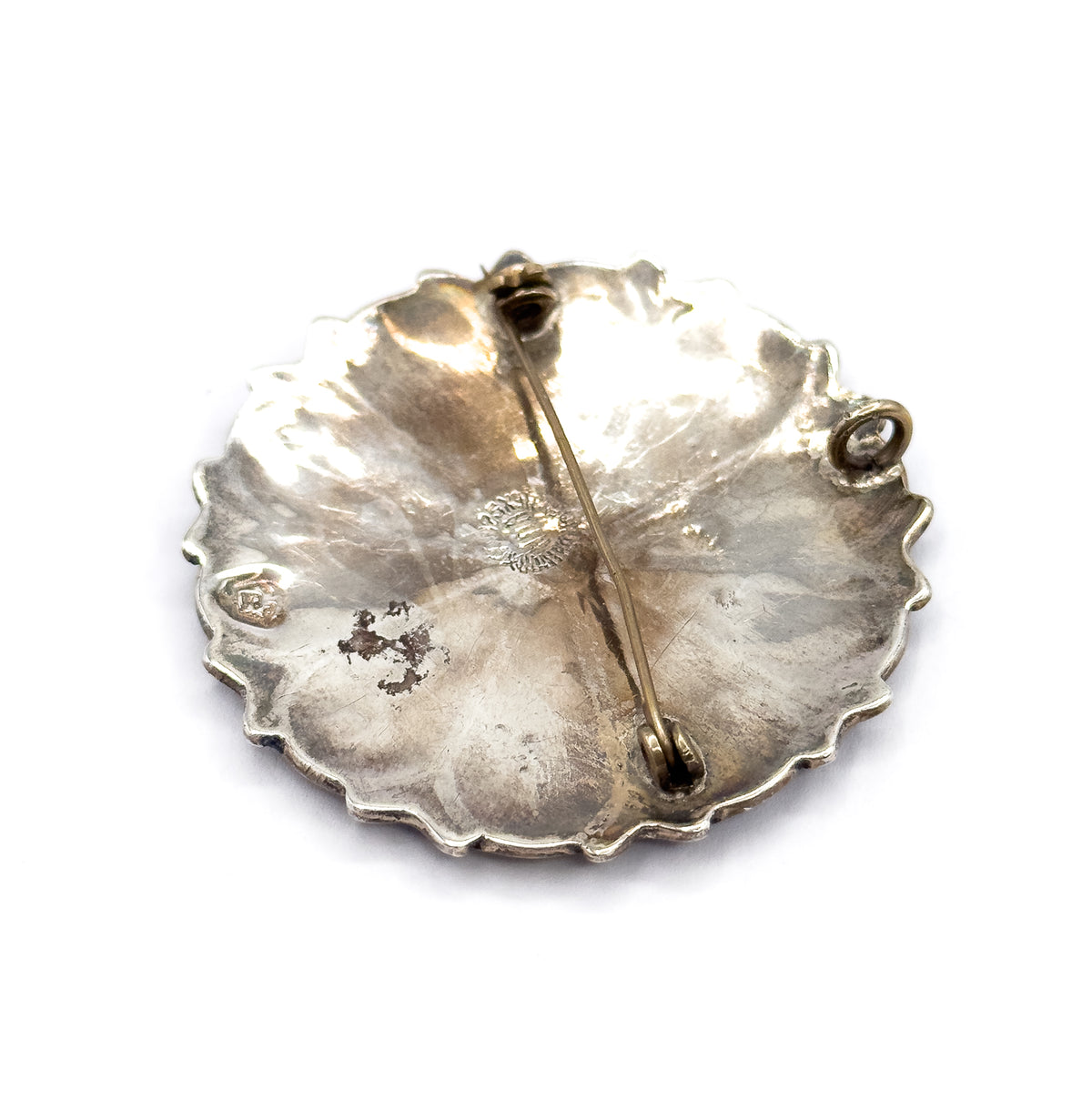 Tilted view of the back on white, the hinge end lifted so the pin stem rises clear of the dished silver, with the open C-shaped catch standing out at the right edge.